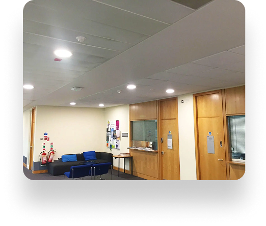 A hallway at Manchester Metropolitan University with white walls and a blue carpet. Blue couches are on the left beneath a noticeboard. On the right, wooden doors with windows line the wall. Emergency equipment is in the corner.