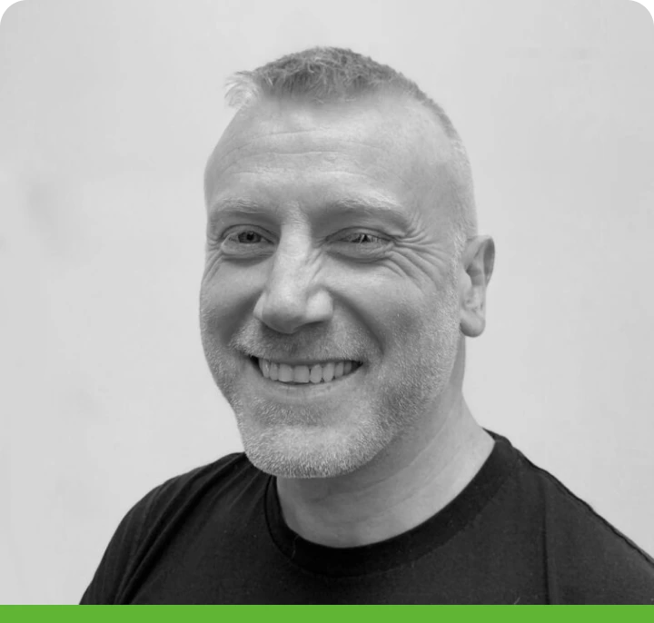 A person with short hair and a beard smiles while wearing a black shirt. The background is plain. The black and white photo has a green border at the bottom.