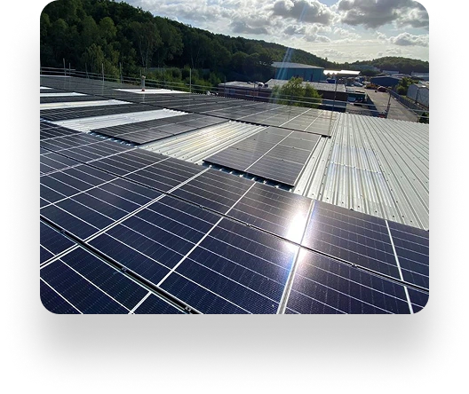Solar panels installed on a large rooftop reflect sunlight. In the background, trees, buildings, and a partly cloudy sky indicate a sunny day.