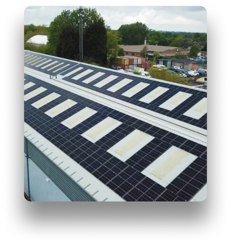 Rooftop view of a long, industrial building fitted with many solar panels. The roof also features white skylights, and trees and other structures are visible in the background.