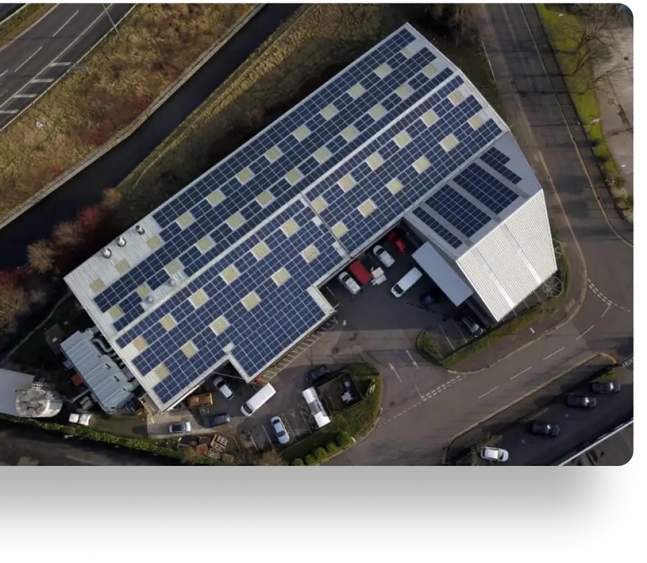 Aerial view of an industrial building with a large array of solar panels on the roof. Several vehicles are parked around the building, and roads are visible on the sides. The surrounding area has some trees and grassy patches.