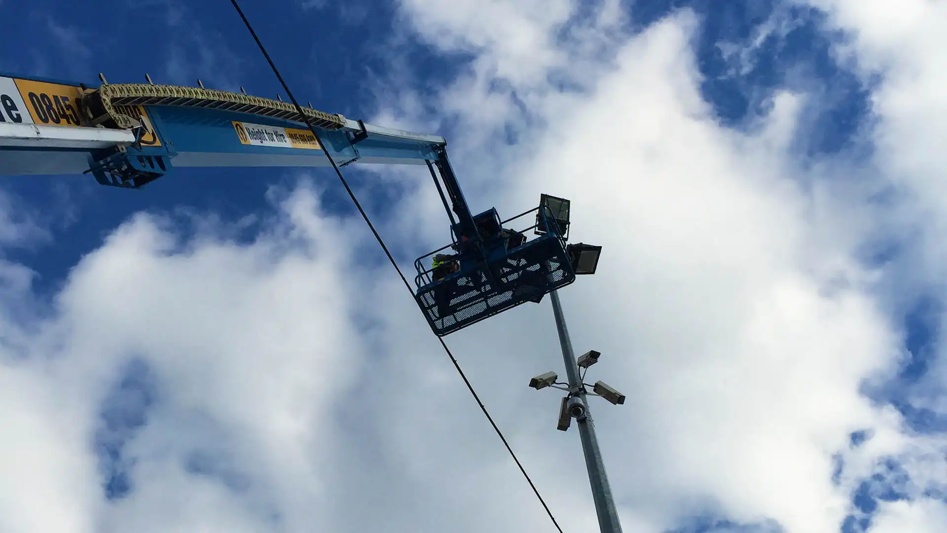 A worker in a blue lift basket, R Swain-style, is elevated near a pole with security cameras. The partly cloudy sky and a power line stretch above.