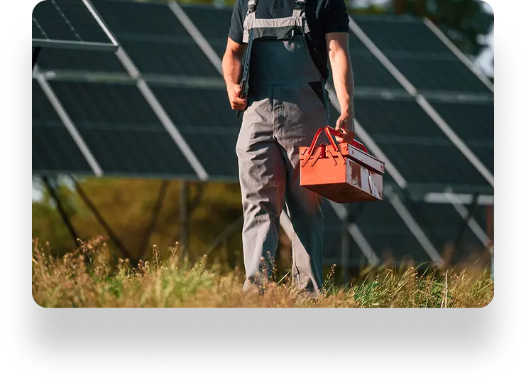 Person in work overalls holding a red toolbox, walking through a grassy area with large solar panels in the background.