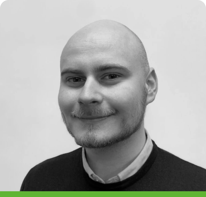 A person with a shaved head and short beard smiles slightly, wearing a dark sweater over a collared shirt. The photo is in black and white with a green border at the bottom.