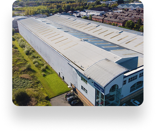 Aerial view of a large industrial warehouse with white roof and skylights, surrounded by greenery and parked cars; residential area in background.