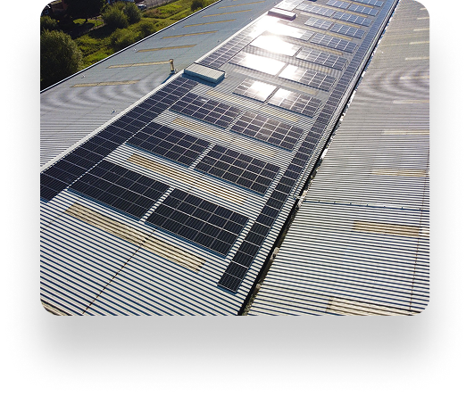 Aerial view of a large industrial roof with Searchlight Electrics reflective solar panels in aligned rows, glinting under sunlight. Nearby is a corrugated metal surface and distant greenery.