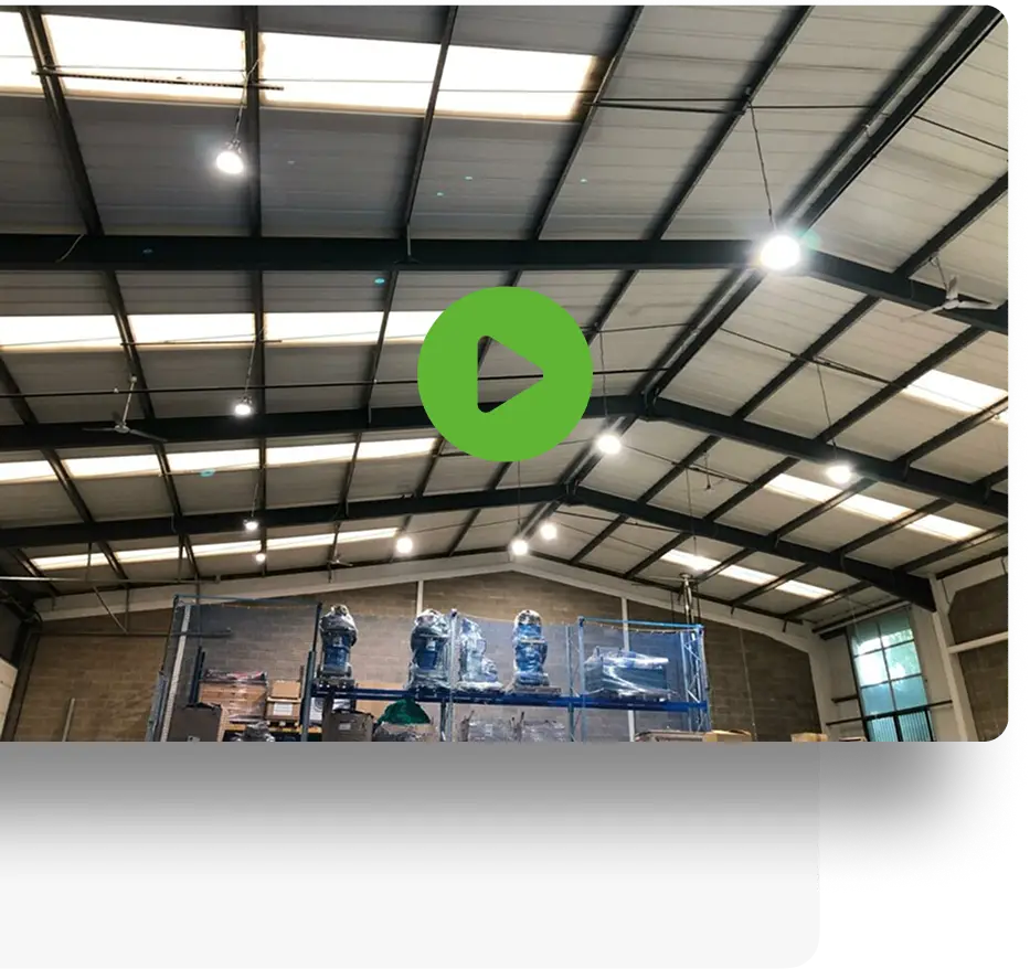 Interior of a spacious warehouse with high ceilings, metal beams, and skylights. A green play button is at the center. Large machines are covered in plastic toward the back of the space.
