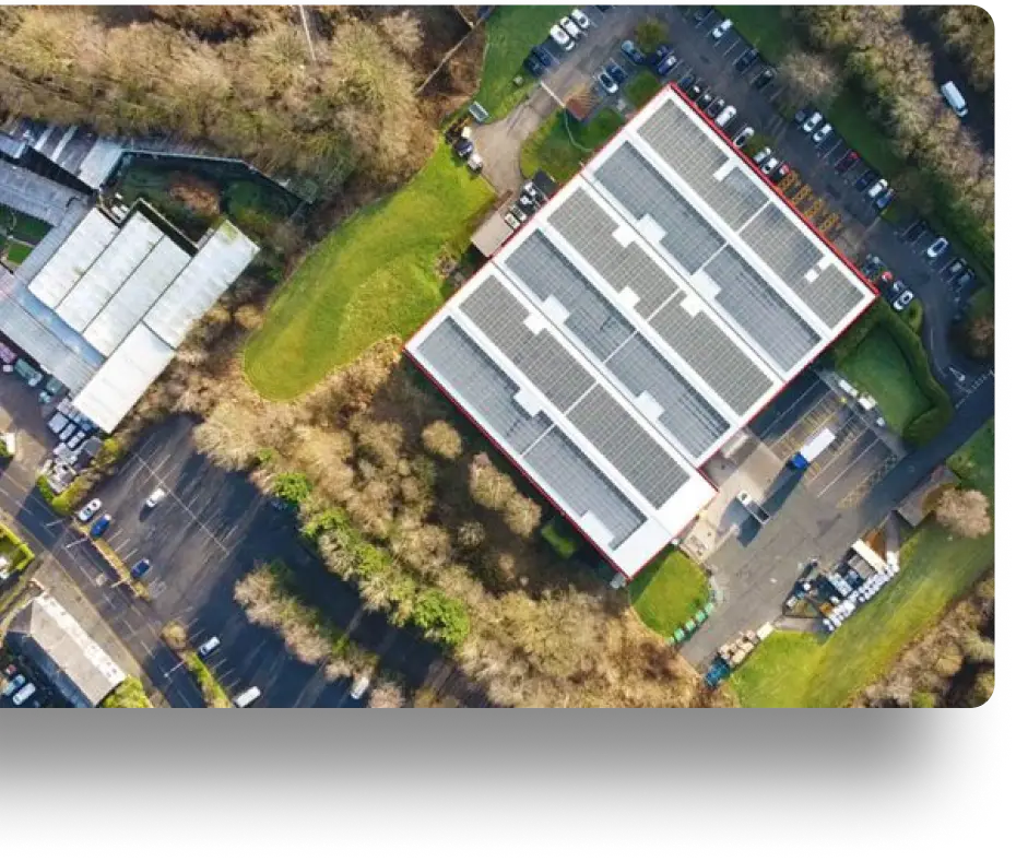 Aerial view of a large rectangular building with a grid-patterned roof surrounded by trees and parking lots. Numerous cars are parked around the building, and there are roads and greenery nearby.