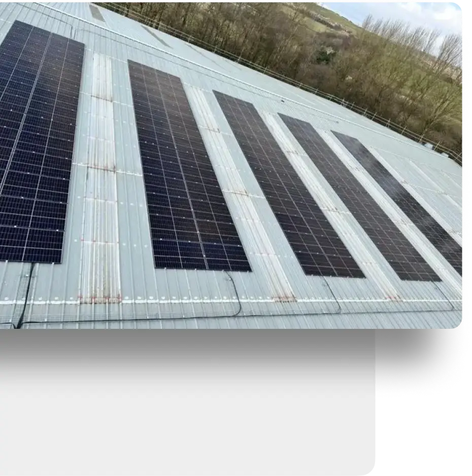 Large solar panels installed on a metal rooftop, set against a backdrop of trees and a hillside. The panels are aligned in rows, maximizing exposure to sunlight. The surrounding area is green and natural.