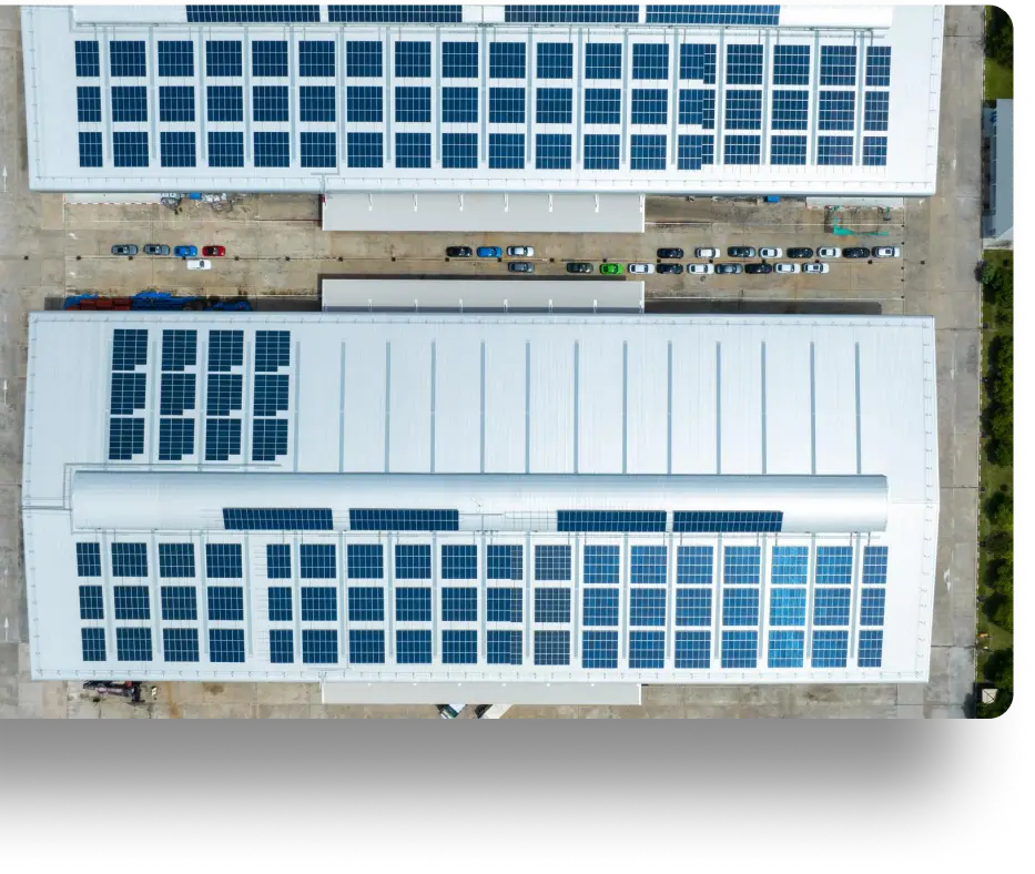 Aerial view of two large white industrial buildings with numerous solar panels on their rooftops. Several parked vehicles are visible between the buildings. Surrounding areas include some greenery and paved surfaces.