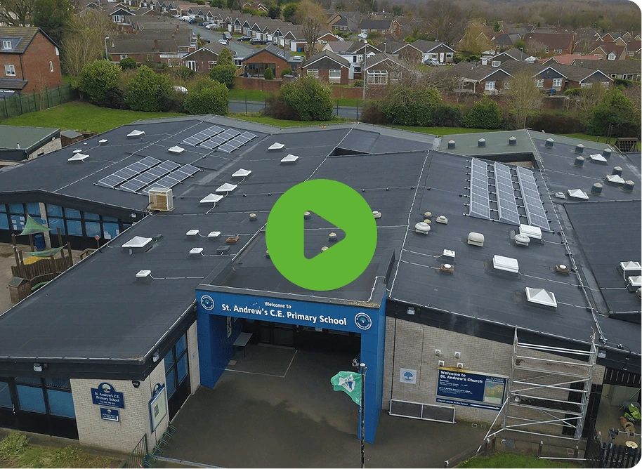 Aerial view of St Andrews Primary School with solar panels on the roof. The entrance sign confirms its St. Andrews C.E. Primary School, surrounded by a neighborhood. A green play button graphic invites exploration.