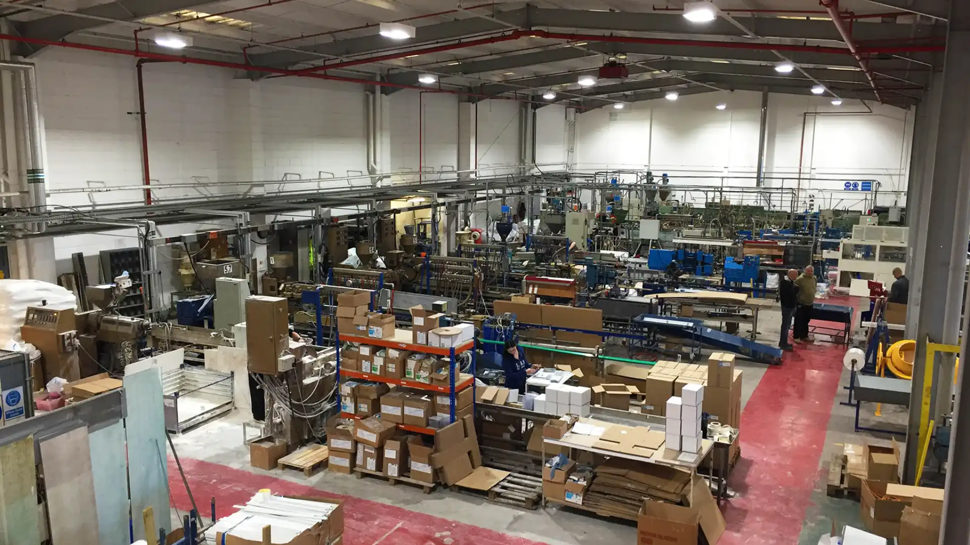 Factory interior with Tatra Rotalac machinery, conveyor belts, and cardboard boxes. Workers are scattered throughout the space. High ceiling has prominent lighting and overhead pipes.