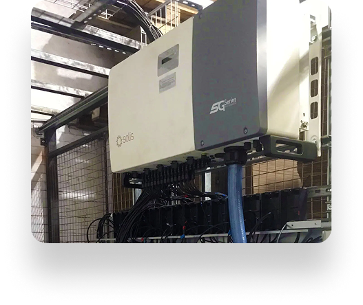 A white Solis SG Series electronic unit with blue and black cables is mounted on a frame at The Box Factory. Metal fencing and structural elements are visible in the background.