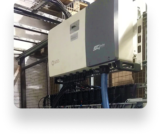 A white Solis SG Series electronic unit with blue and black cables is mounted on a frame at The Box Factory. Metal fencing and structural elements are visible in the background.