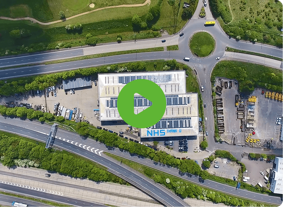 Aerial view of an NHS building with surrounding roads, greenery, and a large roundabout. Vehicles are parked around the site. A green play button icon is centered on the image.