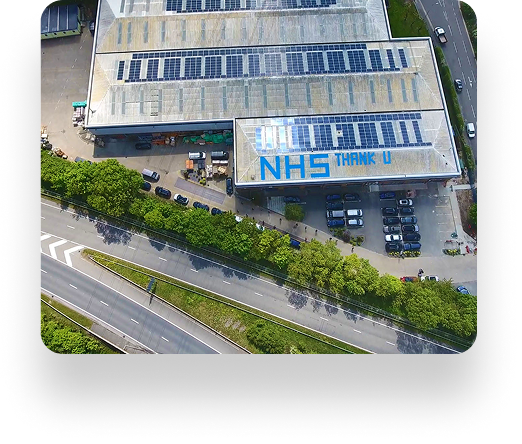 Aerial view of a building with solar panels and large NHS THANK U text on the roof, surrounded by trees, parked cars, a curved intersection, and the Trimline Group facility nearby.