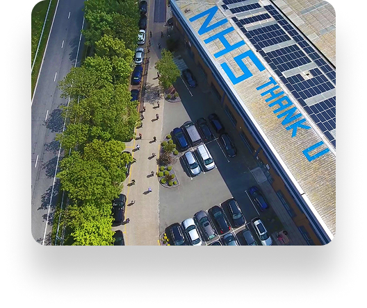 Aerial view of a building roof with NHS THANK U in bold blue by Trimline Group. People stand spaced on the sidewalk below, near green trees and parked cars.