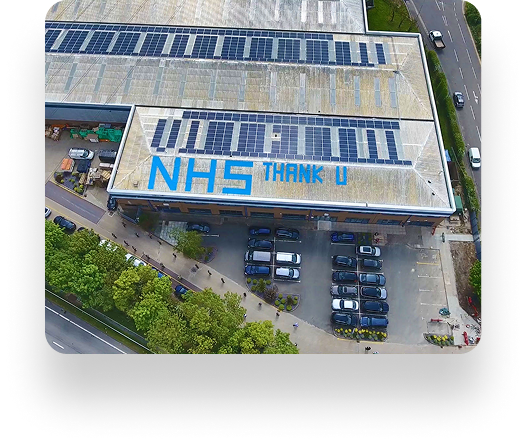Aerial view of a large building with NHS Thank U on the roof, surrounded by solar panels. Cars are in the parking lot, and lush trees encircle the area.