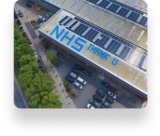 Aerial view of a building with solar panels and NHS THANK U in blue letters on its roof. Trimline Group cars are parked nearby and trees line the sidewalk.