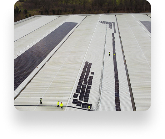TVS Supply Chain Solutions workers in yellow vests install solar panels on a large industrial rooftop, with rows of panels shining under the sun and trees in the background.
