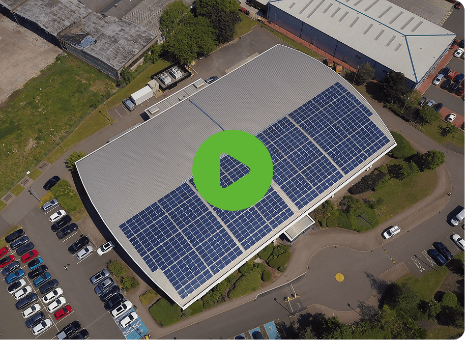 Aerial view of a building with UBT solar panels on the roof, surrounded by parking lots filled with cars. A green play button icon is centered on the image.