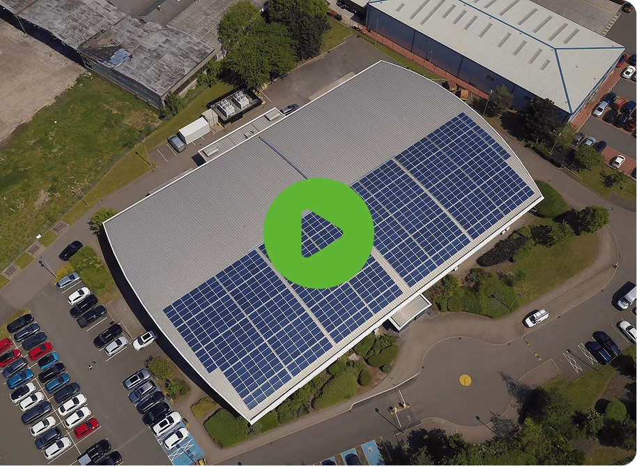 Aerial view of a building with UBT solar panels on the roof, surrounded by parking lots filled with cars. A green play button icon is centered on the image.