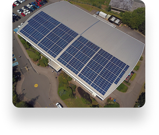 Aerial view of a UBT facility with grey roof and solar panels. Nearby, cars line the road, surrounded by green trees and a small grassy lawn.