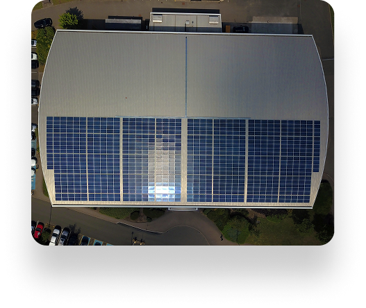 Aerial view of the UBT building with solar panels on its curved roof. Parked cars line the perimeter, surrounded by pathways and lush greenery.