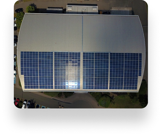 Aerial view of the UBT building with solar panels on its curved roof. Parked cars line the perimeter, surrounded by pathways and lush greenery.
