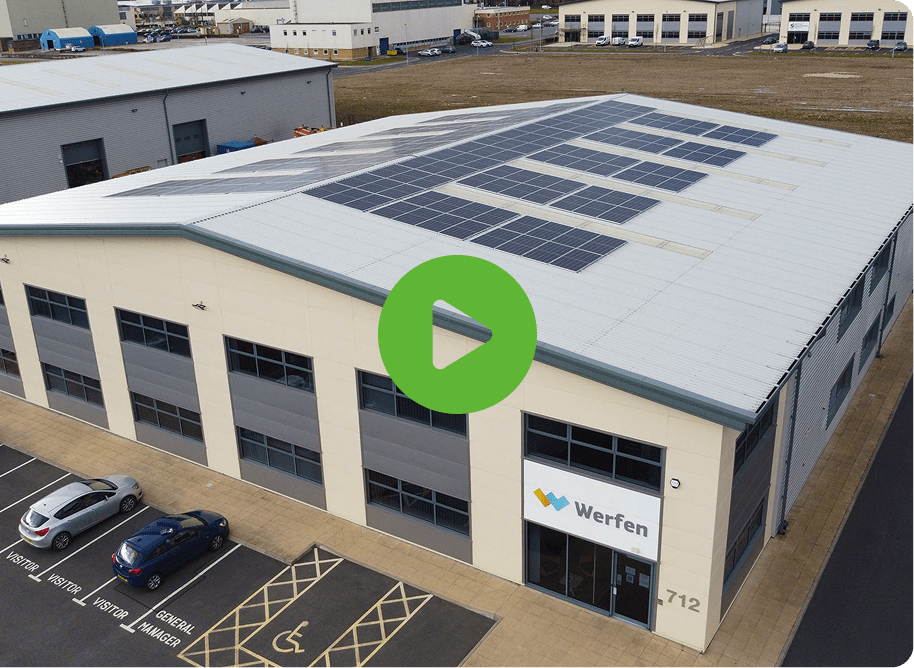 Aerial view of industrial building with solar panels. Wefen on facade, large windows. Cars parked below, one in wheelchair space. Play button overlay.