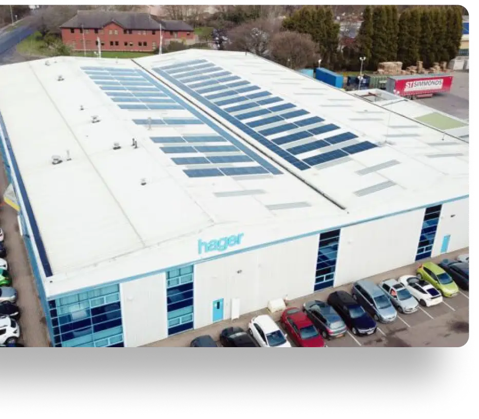 Aerial view of a large industrial building with Hager on the facade. The roof is equipped with numerous solar panels. Several cars are parked in front, and there are trees and another building in the background.