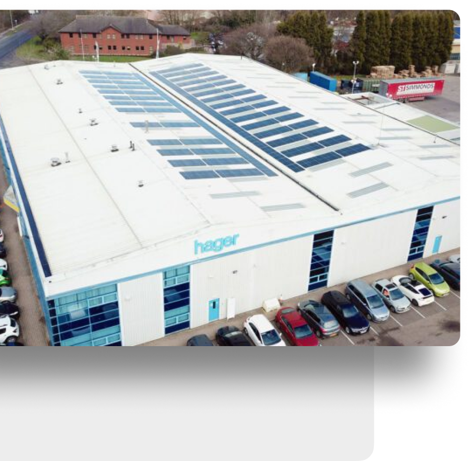Aerial view of industrial building with solar panels on roof, parking lot, and cars nearby.