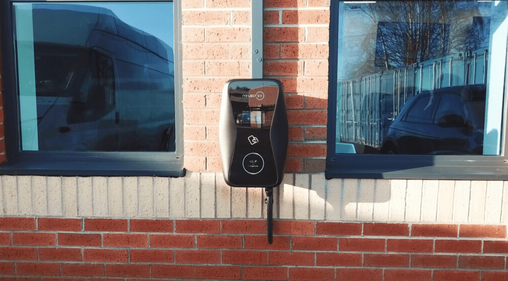 Electric vehicle charging station on a brick wall between two windows, reflecting a van and a car.