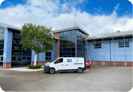 Blue glass building with scaffolding, white van parked in front, red car partially visible, and a tree.