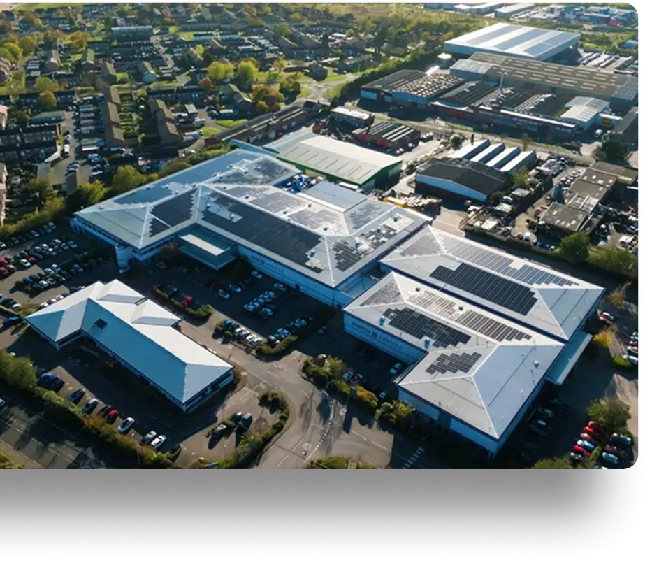 Aerial view of an industrial area with large buildings featuring solar panels. Surroundings include trees, parking lots, residential houses, and other structures.