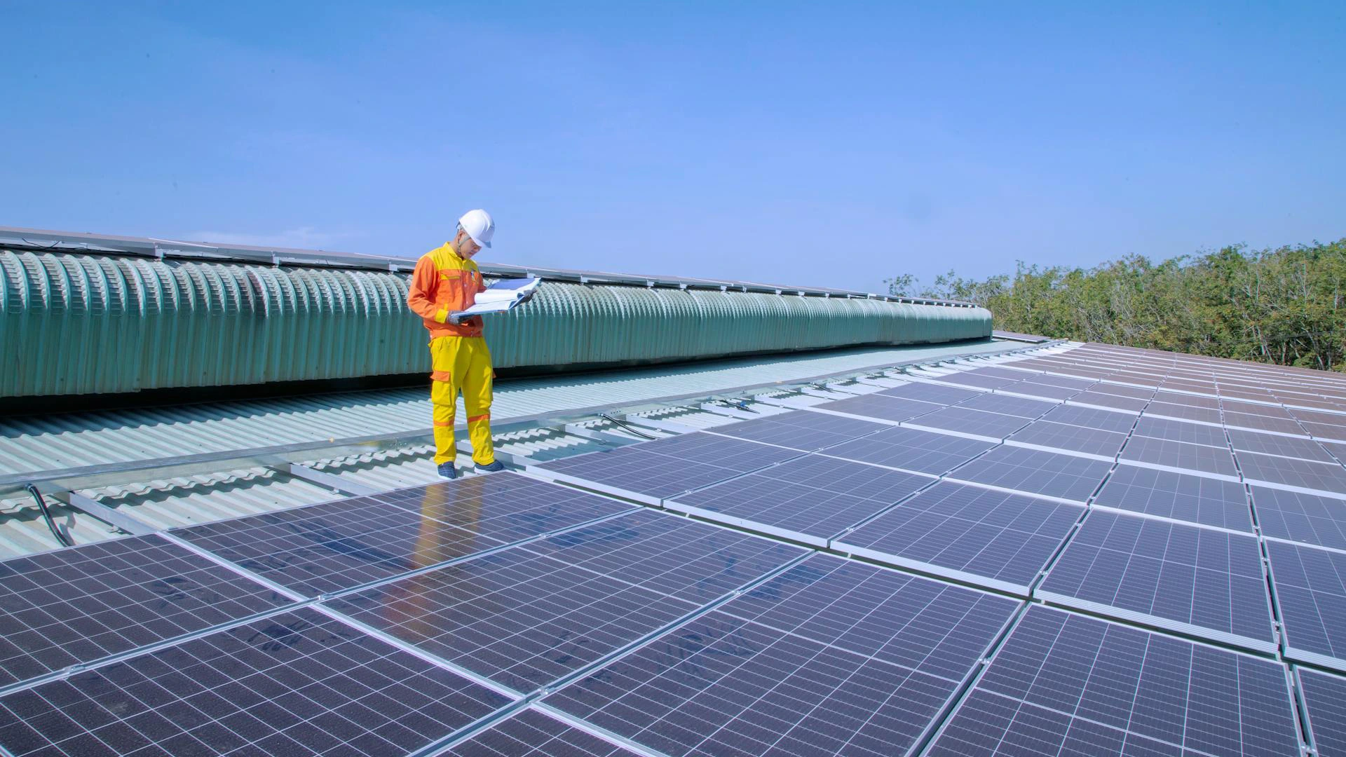 A person in a hard hat and colorful uniform inspects solar panels on a rooftop, holding documents. The sky is clear, with ventilators and trees in the background.