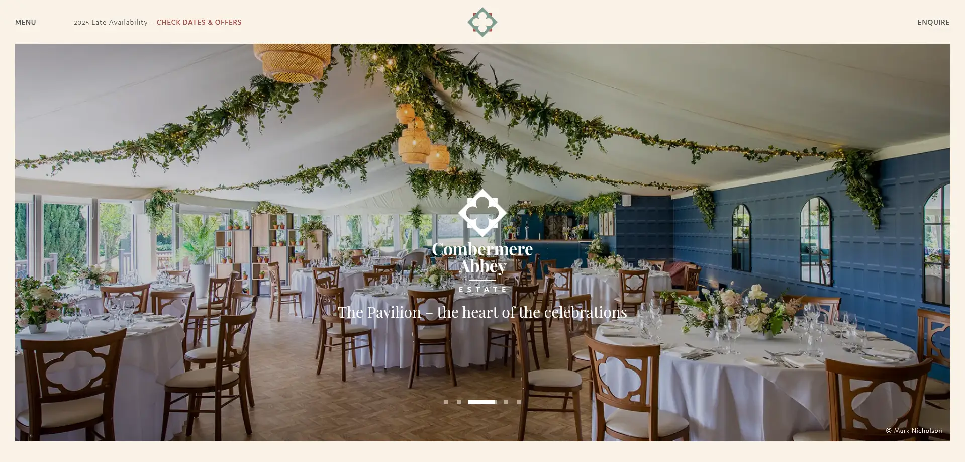 An elegantly decorated event space at The Pavilion, Combermere Abbey Estate, prepared for a formal celebration. The room features round tables draped in white tablecloths, adorned with floral centerpieces. The chairs have a distinctive clover-like pattern on their backrests. The ceiling is decorated with greenery and hanging lights, creating a warm and festive atmosphere. The walls are painted blue and feature large arched mirrors, adding to the sophisticated decor. Natural light streams through sizable windows, offering a view of lush greenery outside.
Overlay text includes:
- "Combermere Abbey Estate"
- "The Pavilion - the heart of the celebrations"
- "2025 Late Availability - CHECK DATES & OFFERS"
- "MENU"
- "ENQUIRE"
- "© Mark Nicholson"
Let me know if you'd like any refinements or a different approach!