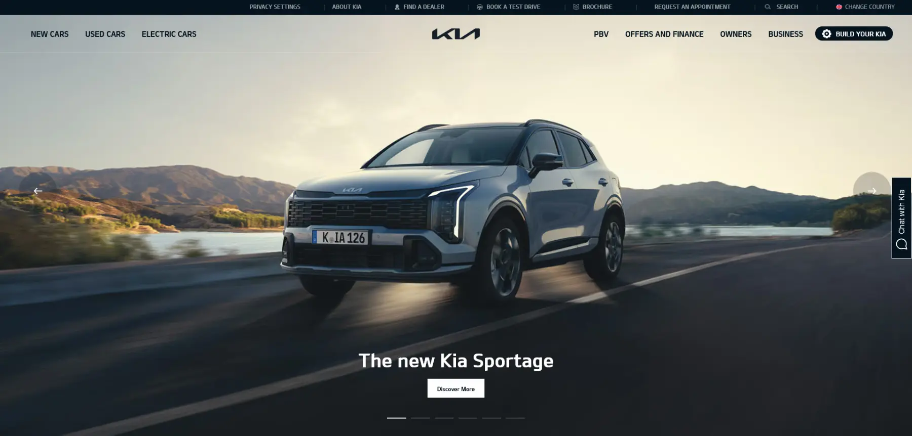 Kia Sportage driving along a scenic coastal road at golden hour with a “The new Kia Sportage” banner and a “Discover More” button.