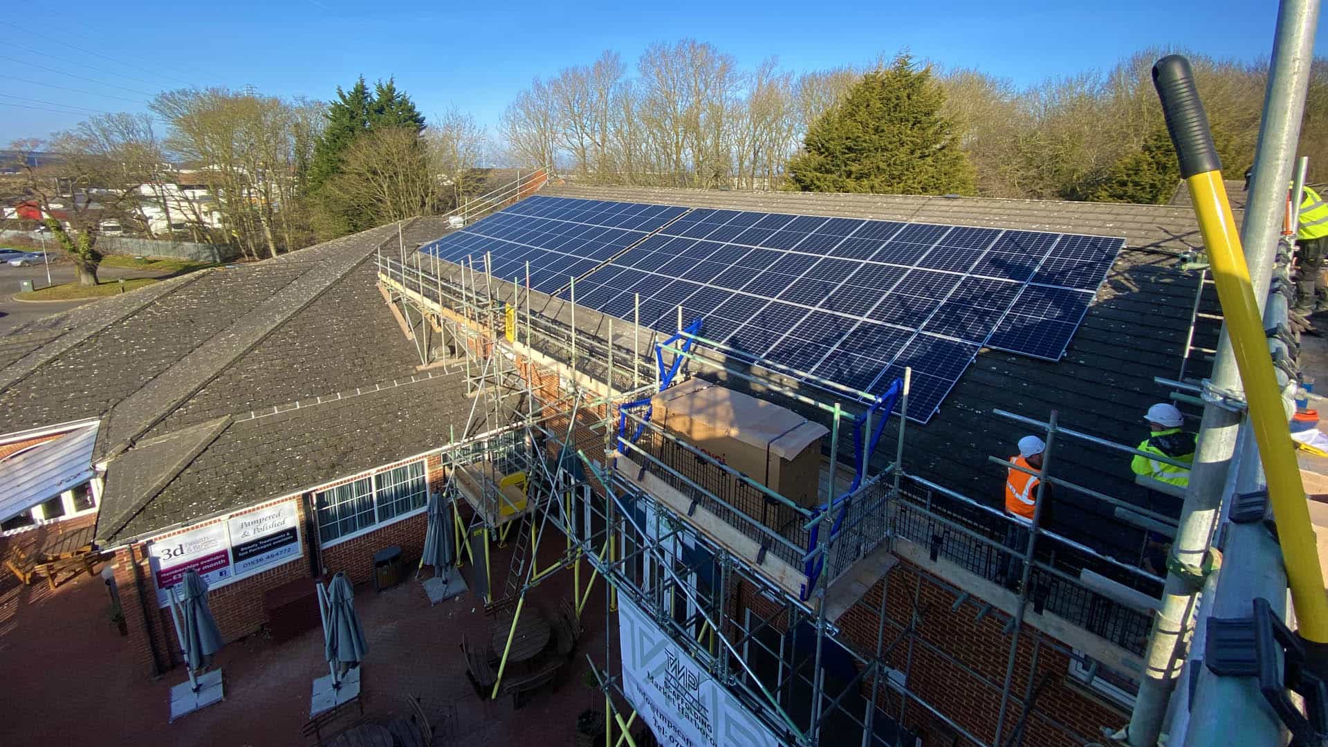 Building with scaffolding, workers in safety gear, and a roof covered in solar panels under clear skies.