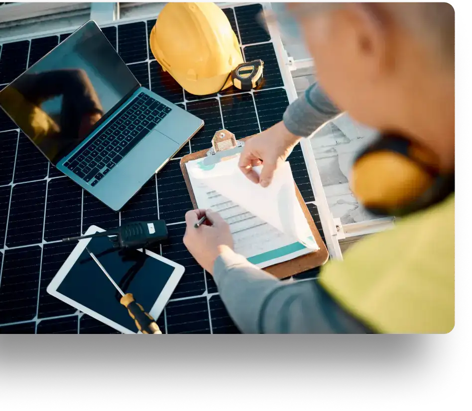 A person in a safety vest is working on a solar panel installation. They hold a clipboard with papers and are writing notes. Nearby are a laptop, a tablet, and a yellow hard hat and safety goggles.
